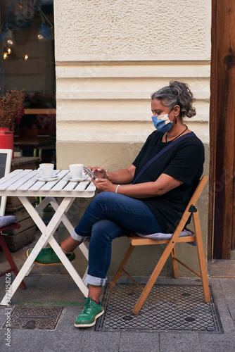 Vertical photo of the side view of a woman with a mask using a smartphone and drinking coffee on the terrace of a bar