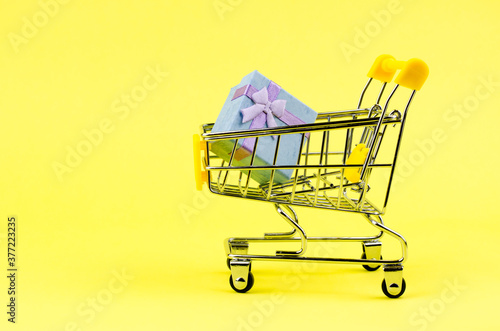 Small gift box in a shopping trolley on a yellow background. Holiday gifts