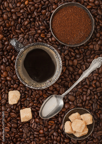 Cup of black coffee with ground coffee and cane sugar with silver spoon inside fresh coffee beans background.