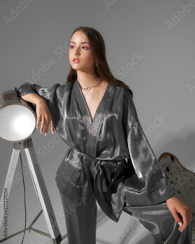 Stylish fashionable girl in silver kimono with trousers standing at white background leaning at lamp. Fashion model with long straight brunette hair and makeup with pink eye shadows studio portrait photo