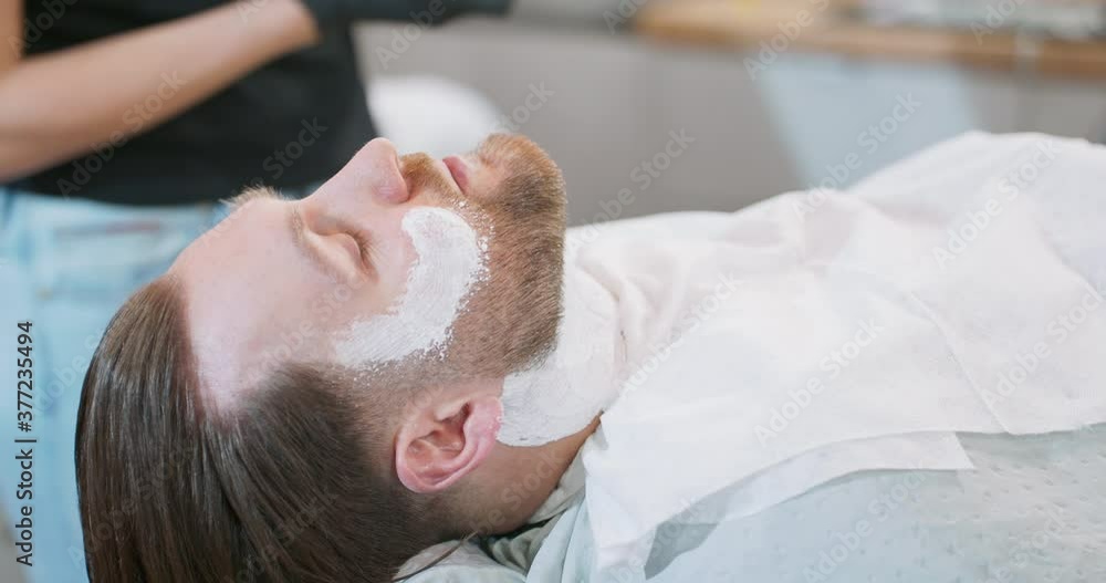 Closeup hands of female barber, who applies with brush shaving cream on young bearded man's face and neck. Closeup hands of female barber, who applies with brush shaving cream on young bearded man's face and neck.