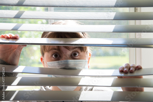 A young woman with face mask looks out from behind blinds 