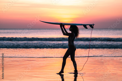 Portrait of woman surfer with beautiful body on the beach with surfboard at colorful sunset in Bali