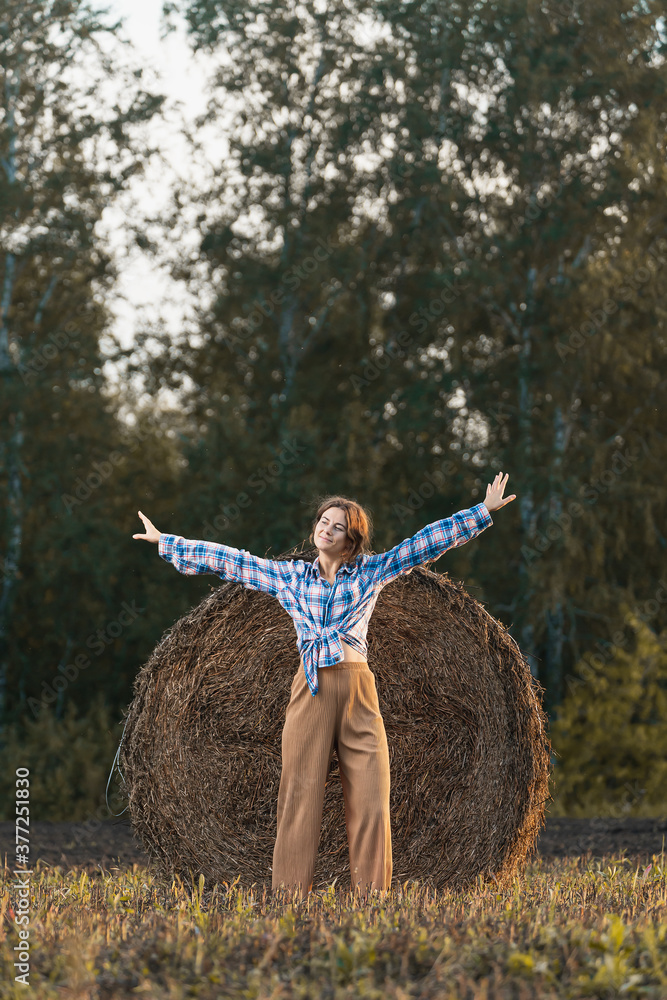 Young beautiful brunette woman in beige trousers and a shirt posing against the background of haystacks in a cut field Young beautiful brunette woman in beige trousers and a shirt posing against the background of haystacks in a cut field