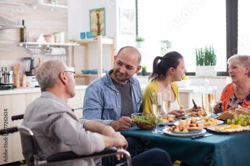 Son looking at disabled senior father in wheelchair during family lunch with healthy food. Happy Mature parents.