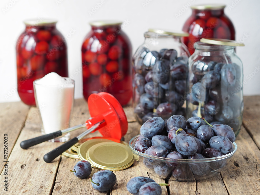 The idea of preserving plum compote for future use in glass jars on a wooden table. Blue plum, capping machine and sugar on the background of finished compote in jars. The idea of preserving plum compote for future use in glass jars on a wooden table. Blue plum, capping machine and sugar on the background of finished compote in jars.