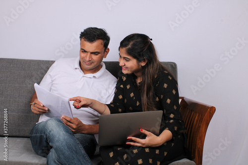 People, money and finances concept. Young Indian woman sitting near her husband on sofa, working with documents and laptop.