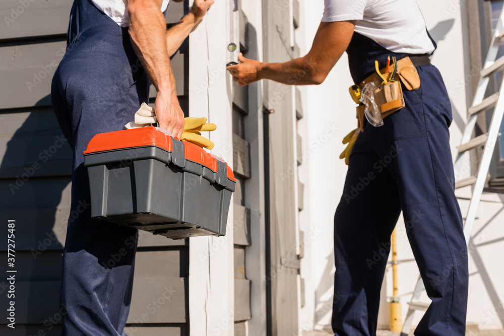 Cropped view of builder holding toolbox and gloves near colleague with spirit level and building Cropped view of builder holding toolbox and gloves near colleague with spirit level and building
