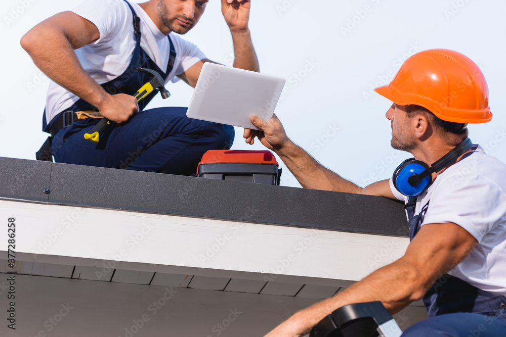 Selective focus of builder showing digital tablet to colleague with hammer on roof of building Selective focus of builder showing digital tablet to colleague with hammer on roof of building
