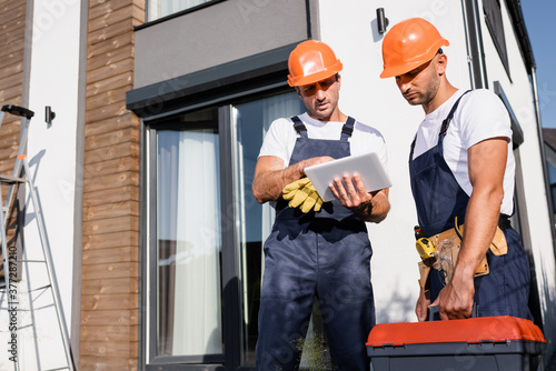 Selective focus of builders in overalls using digital tablet while working near building