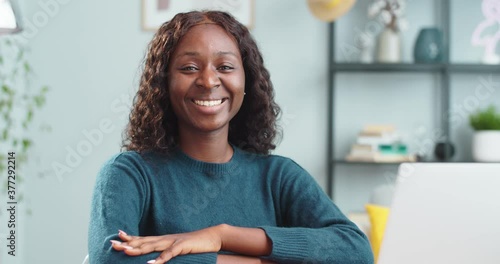 Portrait of joyful pretty African American girl in good mood sitting at desk indoors and smiling. Beautiful cheerful curly female with smile on face looking at camera in room alone. Emotions concept photo