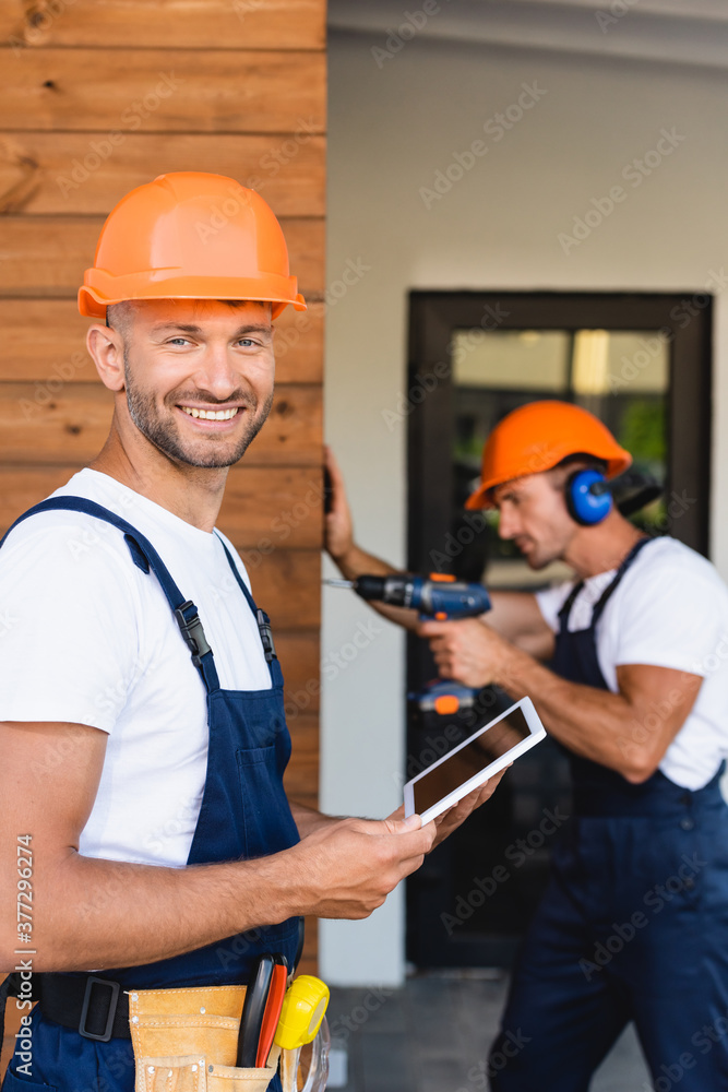 Selective focus of builder holding digital tablet near colleague working with electric screwdriver on facade of building Selective focus of builder holding digital tablet near colleague working with electric screwdriver on facade of building