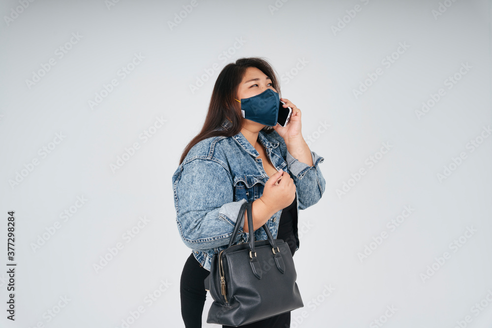 Asian chubby woman wear mask and jeans jacket talking on the phone over white background. Asian chubby woman wear mask and jeans jacket talking on the phone over white background.