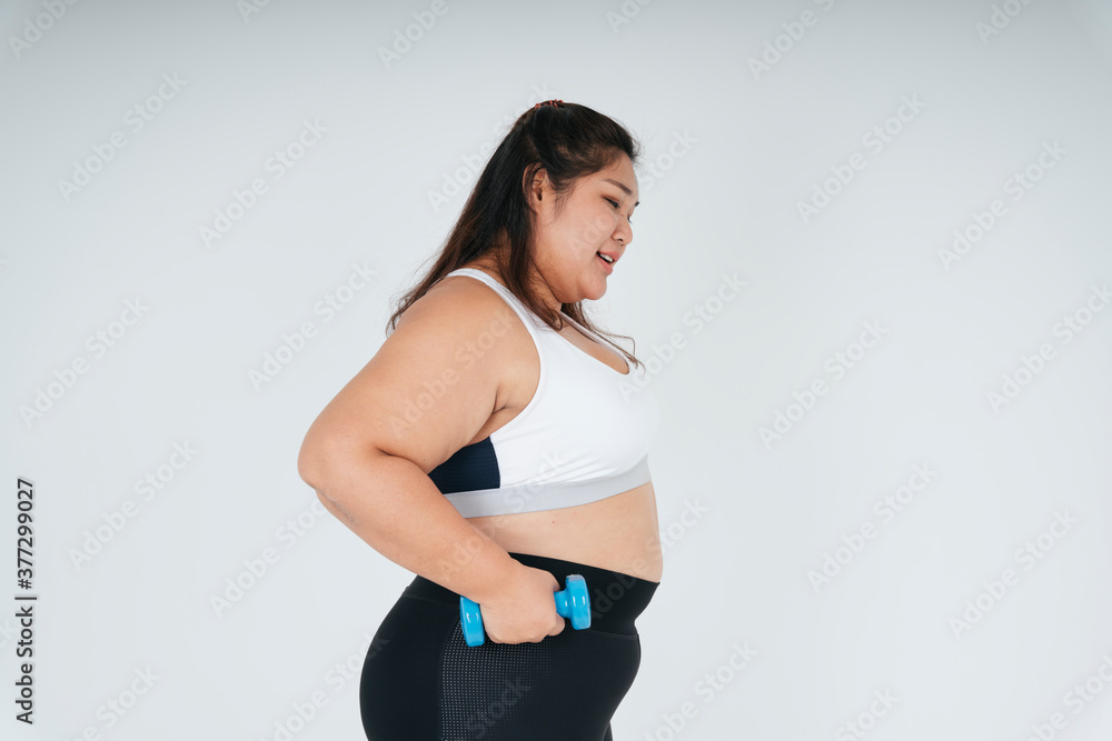 Happy asian chubby woman in white sport bra holding blue dumbbell over white background. Happy asian chubby woman in white sport bra holding blue dumbbell over white background.