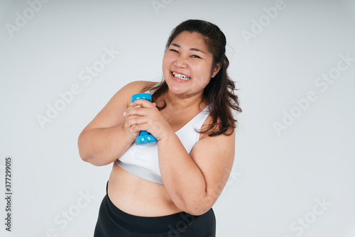 Happy asian chubby woman in white sport bra holding blue dumbbell over white background. © THESHOTS.CO