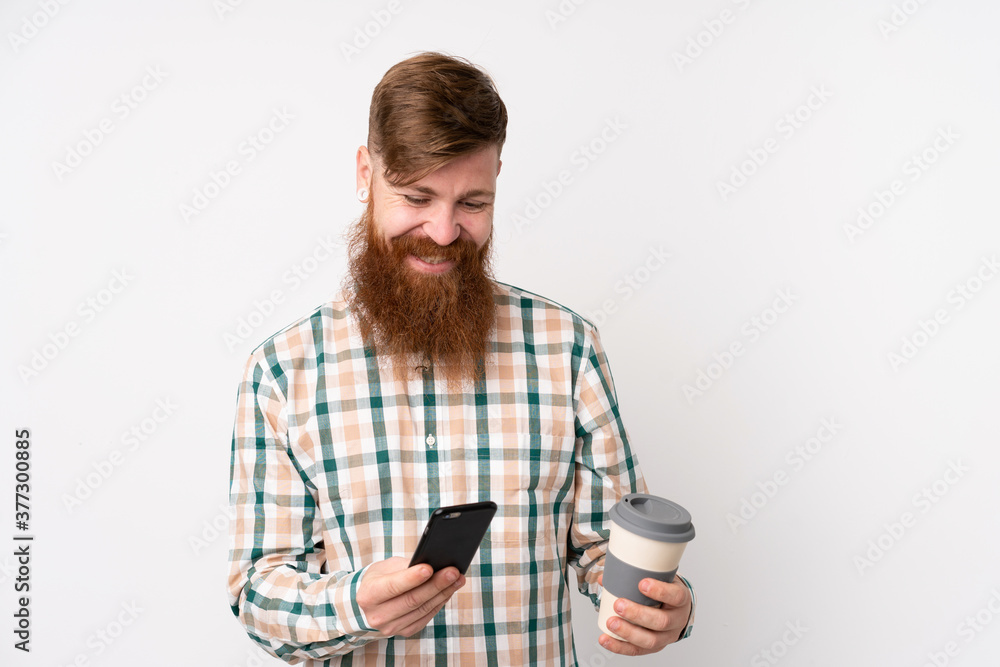 Redhead man with long beard over isolated white background holding coffee to take away and a mobile Redhead man with long beard over isolated white background holding coffee to take away and a mobile