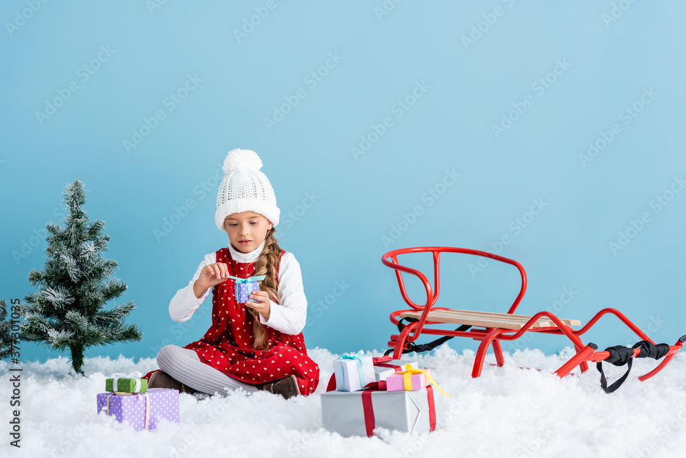 child in hat and winter outfit sitting on snow and holding present near sleigh isolated on blue
