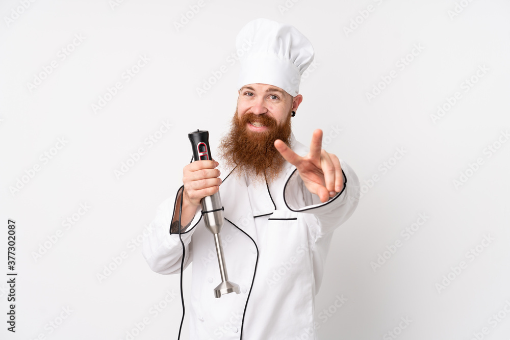 Redhead man using hand blender over isolated white background smiling and showing victory sign Redhead man using hand blender over isolated white background smiling and showing victory sign