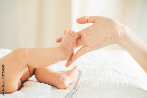 Mother runs fingers on little baby bare feet and toes practicing massage at home against blurry white wall under bright sunlight closeup