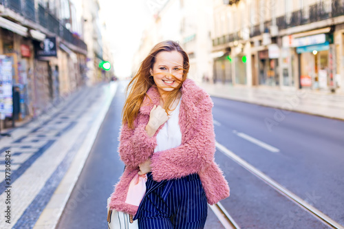 A beautiful girl in a pink fur coat and a white T-shirt is walking along the road on a sunny day. Beautiful young woman with a smile on her face