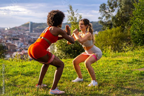 Caucasian blonde girl and dark-skinned girl with afro hair doing squat exercises in a park with the city in the background. Healthy life, fitness, fitness girls, gray and red sport outfits