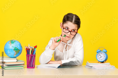 The schoolgirl sits at the desk and thinks about the decision of the task on yellow background. Back to school. The new school year. Child education concept.