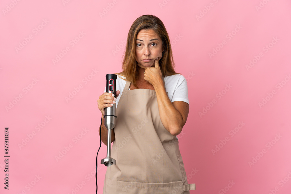 Middle age woman using hand blender isolated on pink background thinking Middle age woman using hand blender isolated on pink background thinking