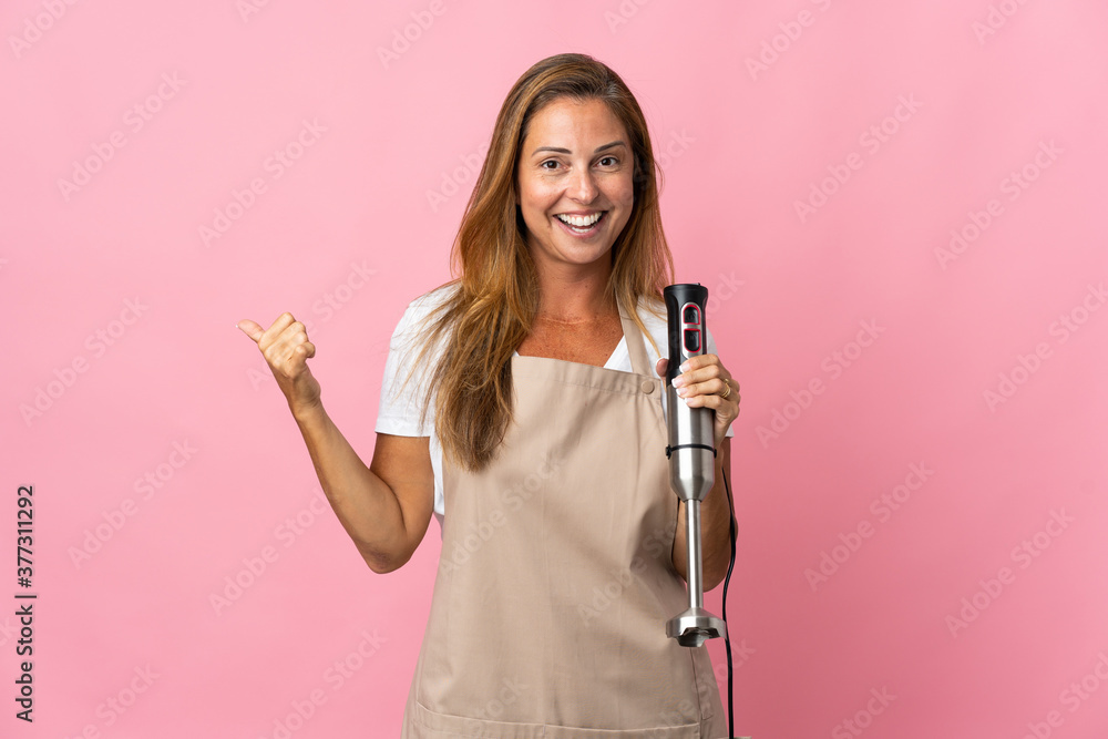Middle age woman using hand blender isolated on pink background pointing to the side to present a product Middle age woman using hand blender isolated on pink background pointing to the side to present a product