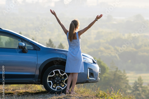 Happy young woman driver with outstretched up hands enjoying warm summer evening standing beside her car. Travelling and vacation concept.