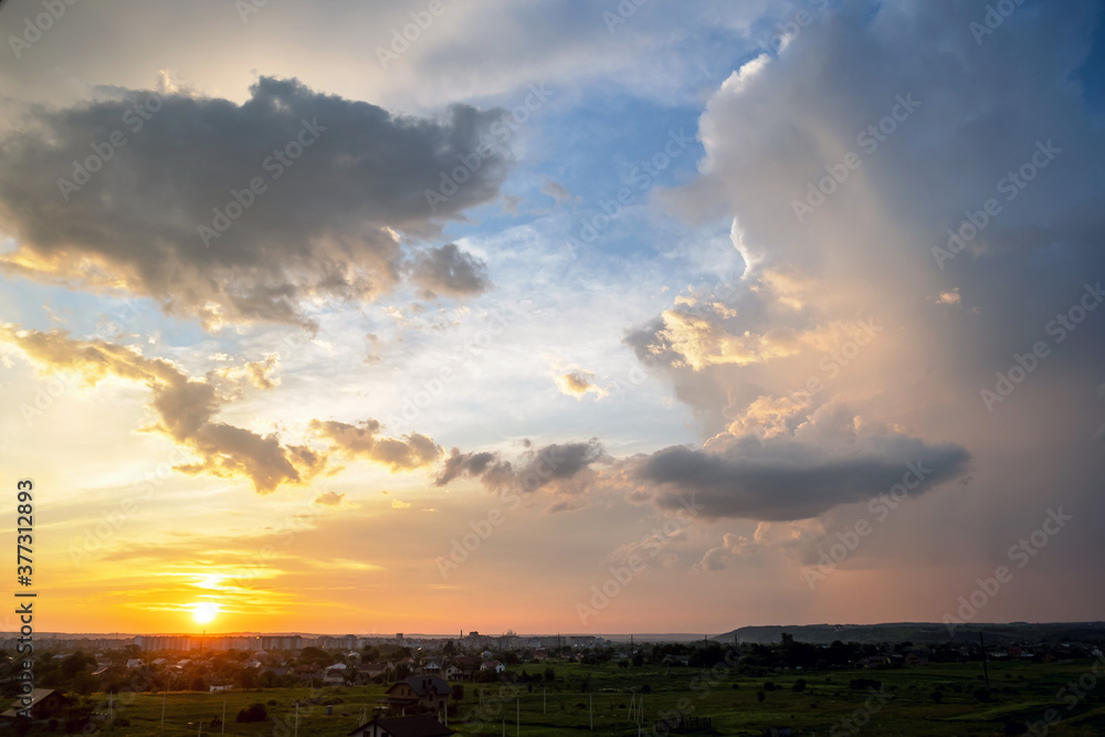 Dramatic sunset landscape of rural area with stormy puffy clouds lit by orange setting sun and blue sky.
