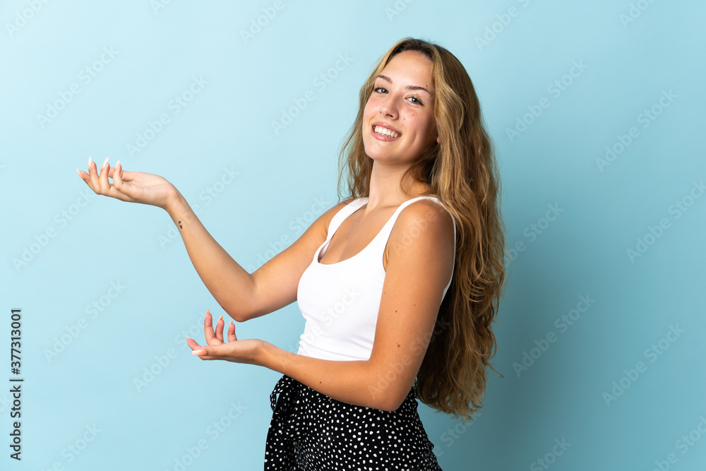 Young blonde woman isolated on blue background extending hands to the side for inviting to come Young blonde woman isolated on blue background extending hands to the side for inviting to come