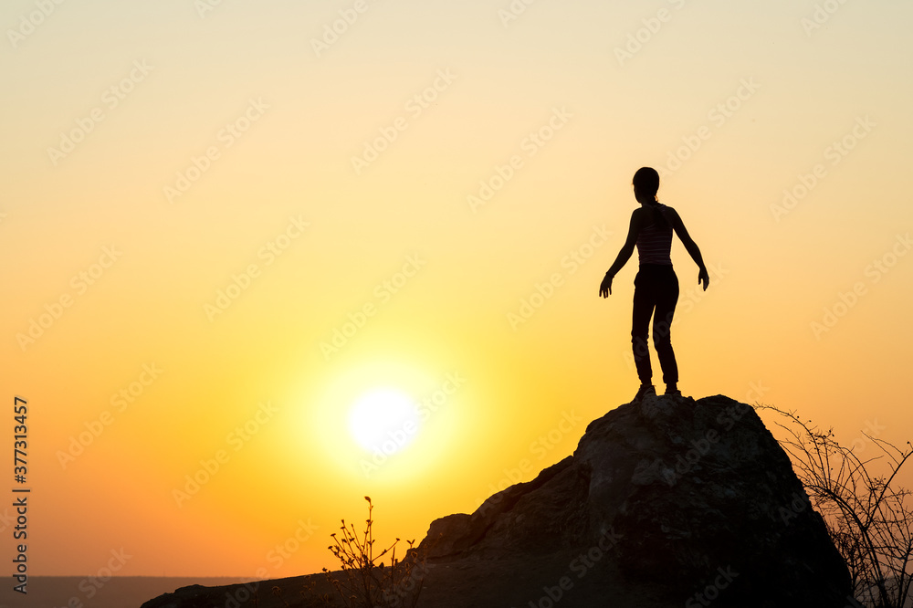 Silhouette of a woman hiker standing alone on big stone at sunset in mountains. Female tourist on high rock in evening nature. Tourism, traveling and healthy lifestyle concept.