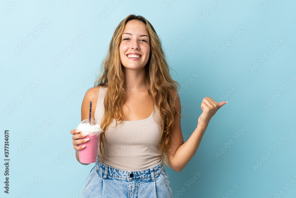 Young woman with strawberry milkshake isolated on blue background pointing to the side to present a product Young woman with strawberry milkshake isolated on blue background pointing to the side to present a product