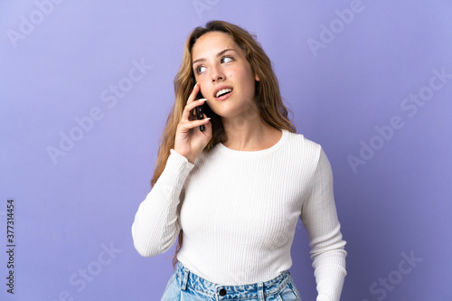Young blonde woman isolated on purple background keeping a conversation with the mobile phone with someone