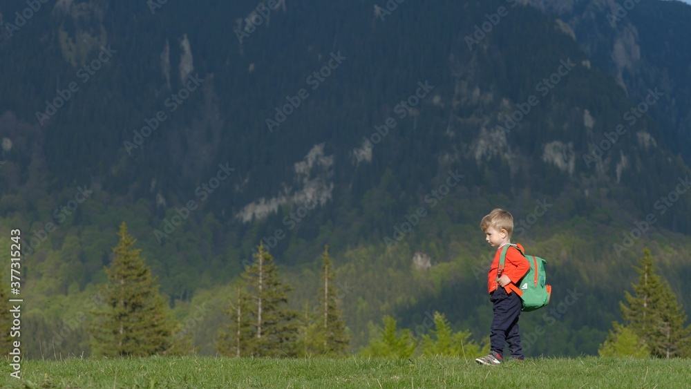 Funny child with backpack walking on green hill in mountains, little toddler hiking
