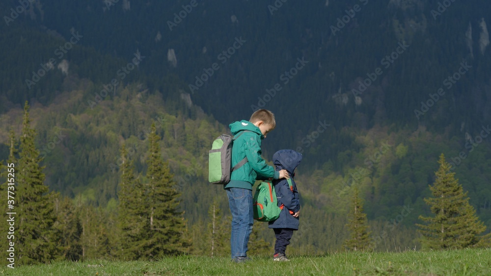 Brother help little child to arrange backpack, friends go together in mountains trip