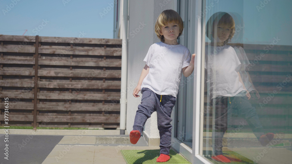 Little blond hair child standing in front of house door going inside, sunny day