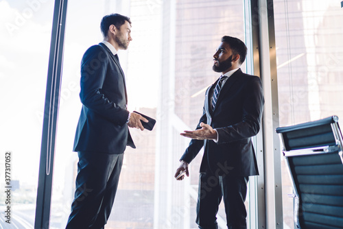 Diverse colleagues dressed in elegant suit communicate near panoramic window in corporate office, multiracial male partners 30 years old discussing productive collaboration and brainstorming briefing