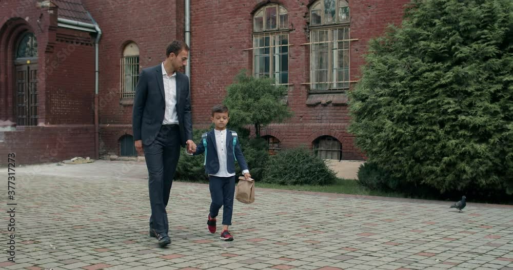 Handsome dad businessman and litlle child with bag talking while holding hand in hand. Man with his cute son walking at school yard. Concept of education and children. Handsome dad businessman and litlle child with bag talking while holding hand in hand. Man with his cute son walking at school yard. Concept of education and children.