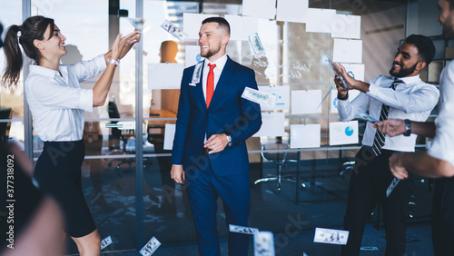 Cheerful male corporate director in formal suit standing in center of room and enjoying monetary rain with happy employees holding dollars in hand and tossing up, concept of wealthy and abundance