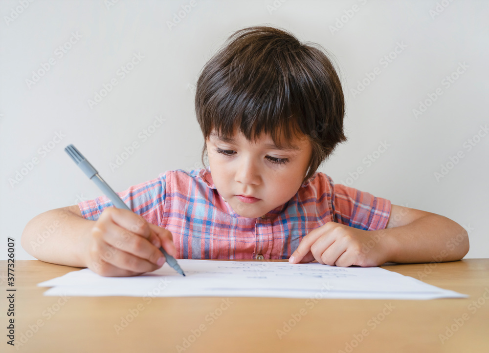 Portrait of school kid boy sitting alone doing homework, Child holding grey colouring on paper, A boy drawing and writing on white paper on table, Elementary school and homeschooling concept Portrait of school kid boy sitting alone doing homework, Child holding grey colouring on paper, A boy drawing and writing on white paper on table, Elementary school and homeschooling concept