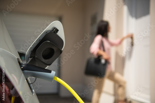 Close up of a electric car charger with female silhouette in the background, entering the home door