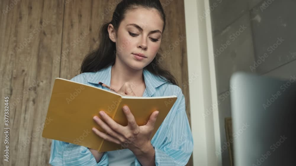 A focused young woman is writing something looking to the camera in her notebook sitting at home A focused young woman is writing something looking to the camera in her notebook sitting at home