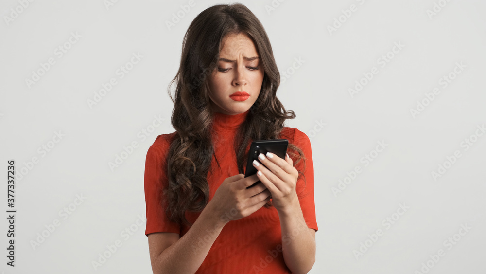 Upset offended girl in red top disappointedly using smartphone over white background Upset offended girl in red top disappointedly using smartphone over white background