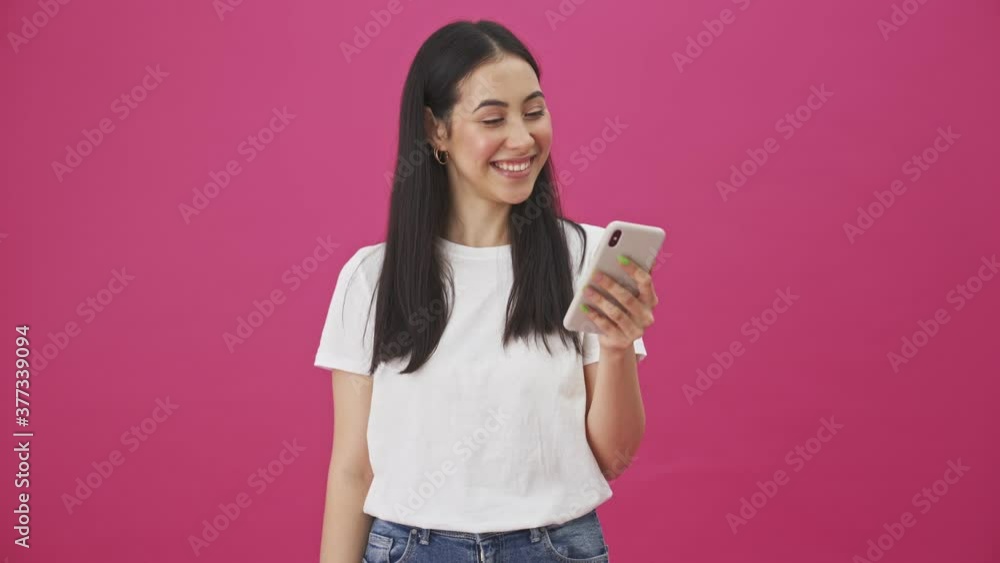 A smiling woman is looking to her smartphone and doing thumb-up gesture standing isolated over a pink background A smiling woman is looking to her smartphone and doing thumb-up gesture standing isolated over a pink background