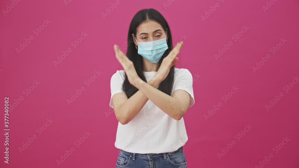 A pretty young woman with protective mask is showing stop gesture while crossing her arms standing isolated over a pink background A pretty young woman with protective mask is showing stop gesture while crossing her arms standing isolated over a pink background