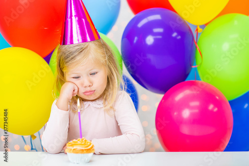 Sad little girl wearing party's cap sits with birthday cupcake