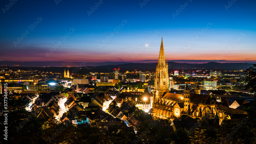 Germany, Freiburg im Breisgau, Magical blue hour atmoshpere of the illuminated skyline and minster cathedral, aerial view above with moon