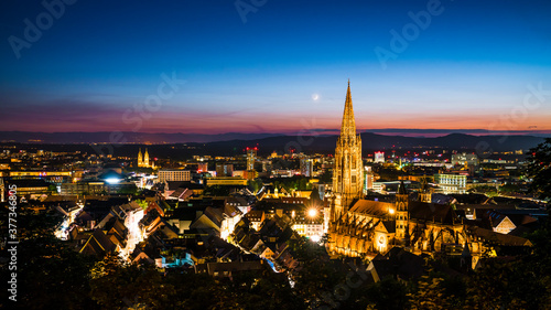 Germany, Freiburg im Breisgau, Magical blue hour atmoshpere of the illuminated skyline and minster cathedral, aerial view above with moon