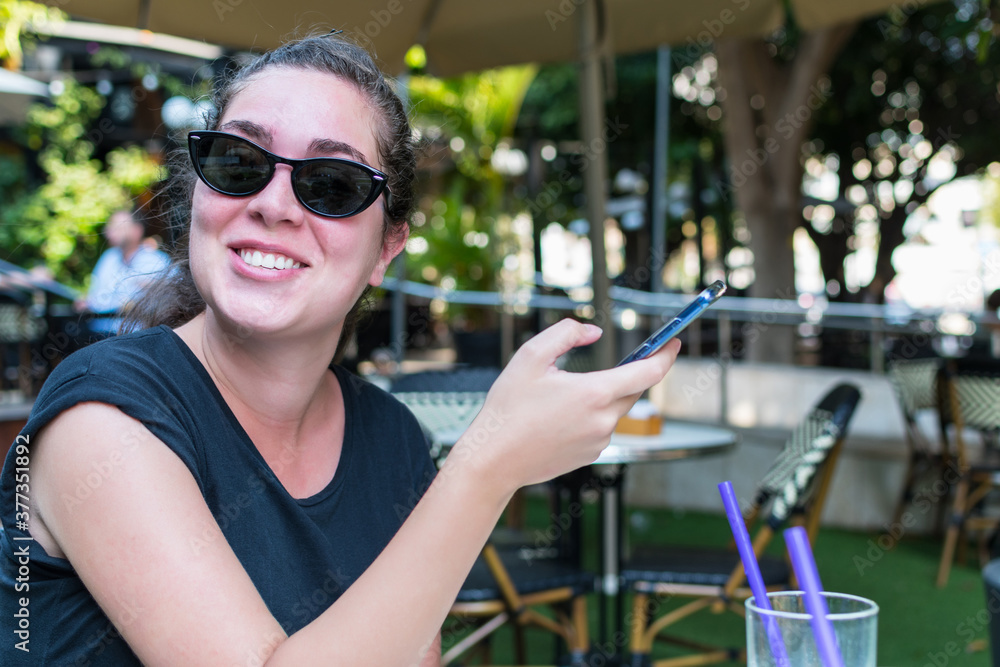 Young woman in sunglasses, laughs happily while checking her cell phone, sitting in an outdoor bar.